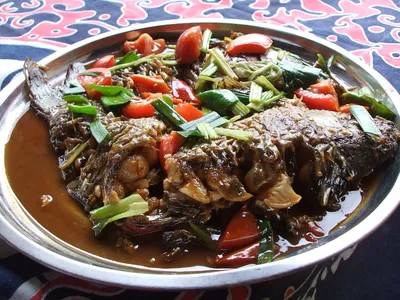 A whole braised beer fish topped with tomatoes, chilies, and scallions on a large plate