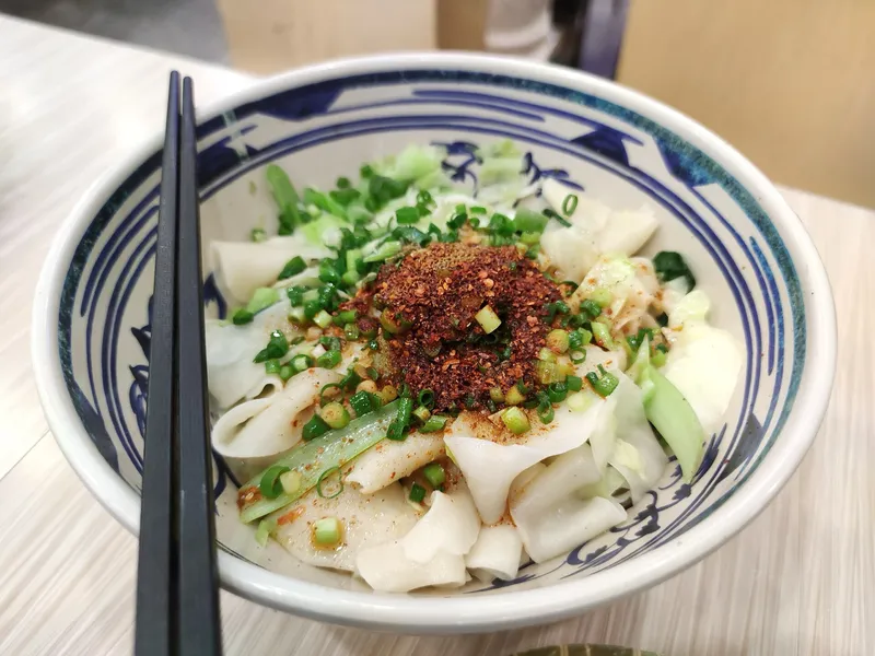 A bowl of wide hand-pulled noodles topped with chili flakes and scallion