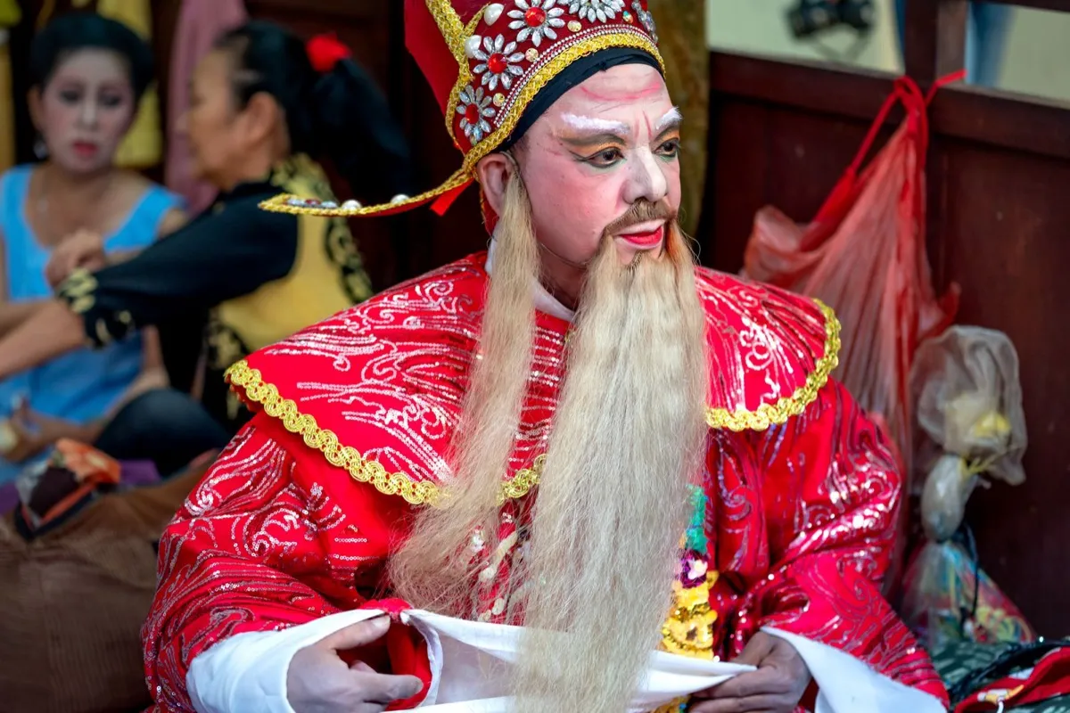 A Chinese opera performer in ornate red costume and white face paint backstage