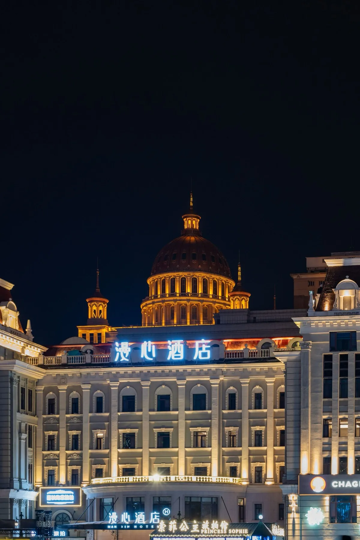 An ornate domed building glowing with warm lights above street-level shops on Harbin's Central Street at night