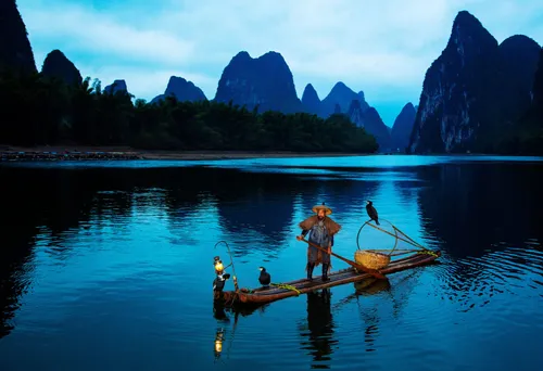 A cormorant fisherman standing on a bamboo raft with lantern and trained birds at dusk on the Li River, karst peaks behind
