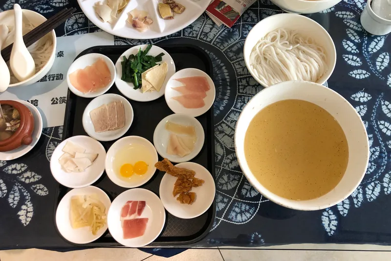 A large bowl of golden broth beside small plates of raw meat, herbs, and rice noodles for crossing-the-bridge noodles