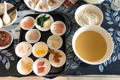 A large bowl of golden broth beside small plates of raw meat, herbs, and rice noodles for crossing-the-bridge noodles