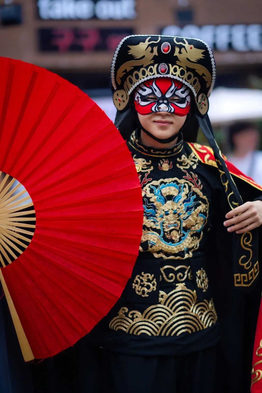A Chinese opera performer in an ornate painted mask holding a red fan
