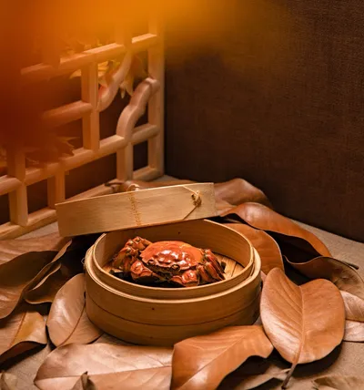 A whole steamed hairy crab in a bamboo steamer basket surrounded by autumn leaves