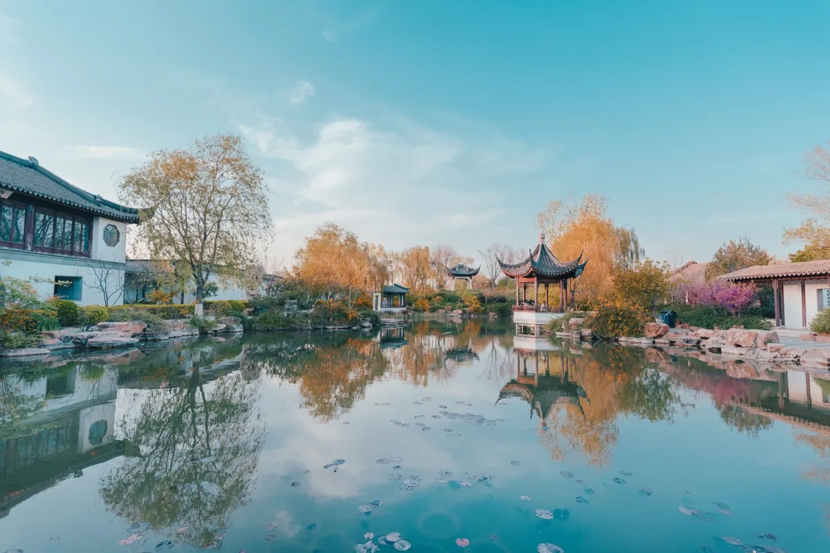 A pavilion beside a lily-covered pond reflecting autumn trees in a classical Suzhou garden