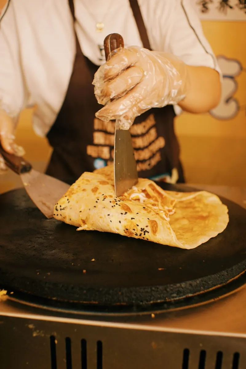 A street vendor folding a sesame-flecked jianbing crêpe on a round griddle