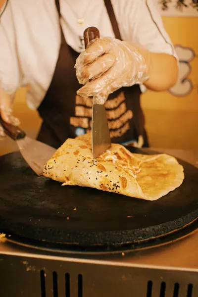 A street vendor folding a sesame-flecked jianbing crêpe on a round griddle