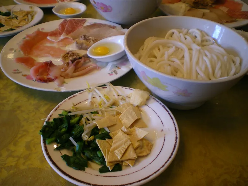 A crossing-the-bridge noodles spread in Kunming: a bowl of hot broth, raw meat slices, egg yolks, and rice noodles on separate plates