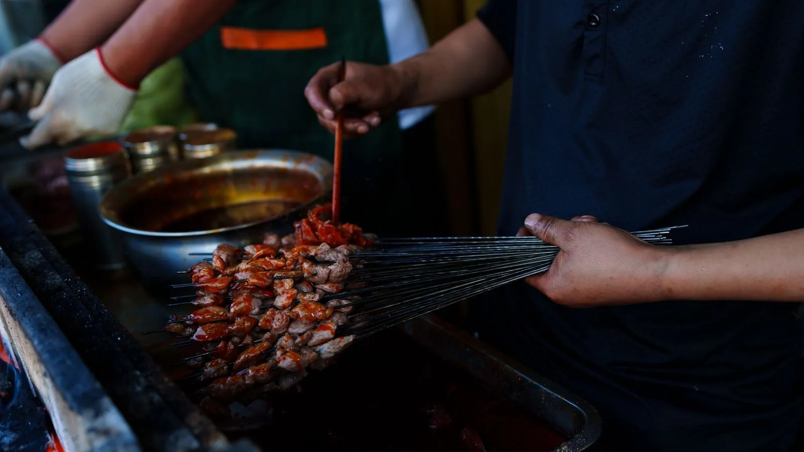 A street vendor grilling and seasoning bundles of lamb skewers over charcoal