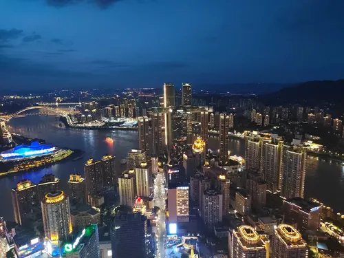 Aerial night view of Chongqing's illuminated skyscrapers and bridges at the confluence of the Yangtze and Jialing rivers, with a lit cruise boat on the water