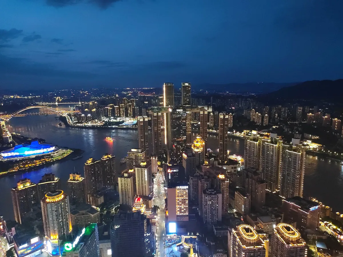Aerial night view of Chongqing's illuminated skyscrapers and bridges at the confluence of the Yangtze and Jialing rivers, with a lit cruise boat on the water