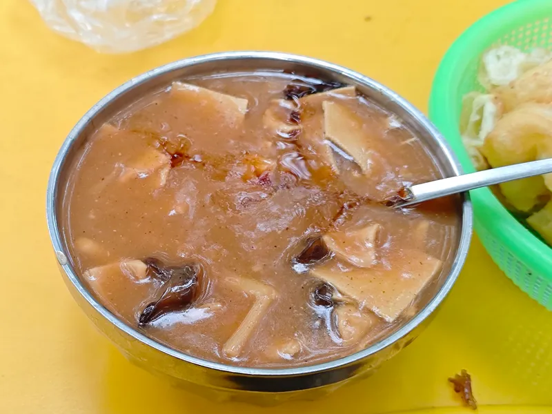A metal bowl of thick, brown hu la tang pepper soup with tofu skin and mushroom, served beside fried dough sticks