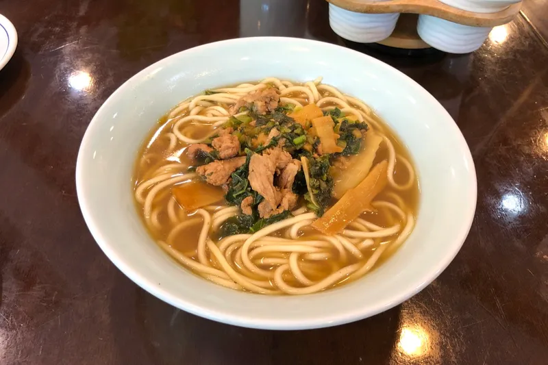 A bowl of pian er chuan noodle soup topped with pork, bamboo shoot, and pickled greens