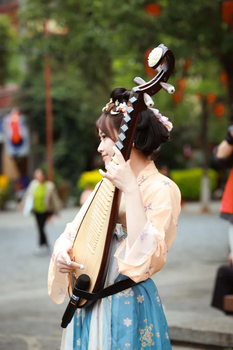 A performer in traditional dress playing a pipa lute outdoors near red lanterns