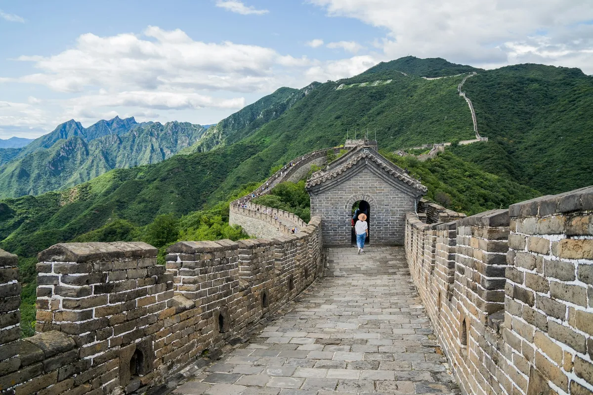 A solo traveler in a sun hat walking through a watchtower archway on the Great Wall, mountains stretching behind