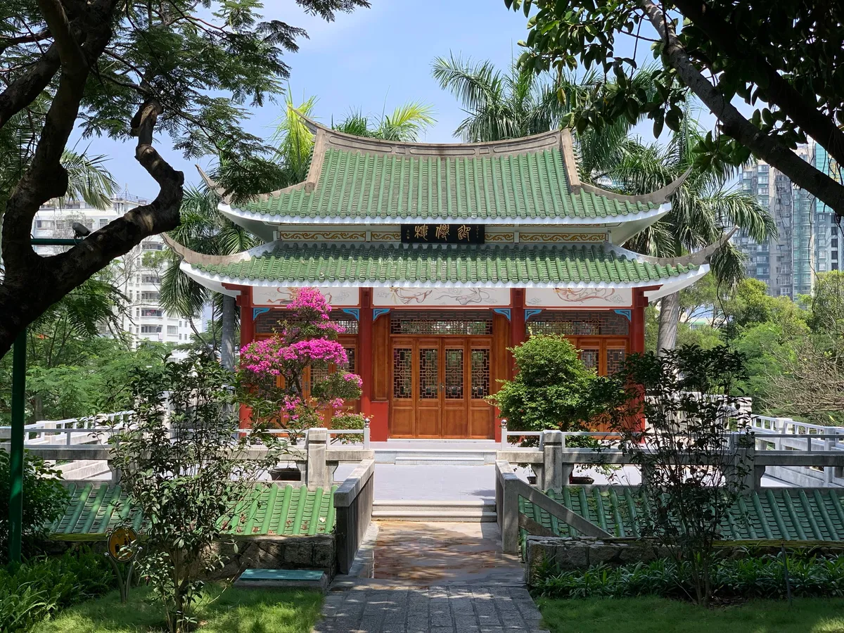 A green-roofed pavilion with red doors and hanging lanterns at South Putuo Temple, framed by palm trees