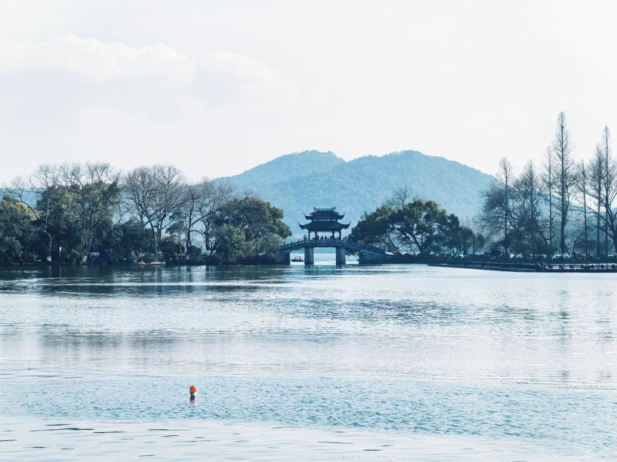 A traditional pavilion on a stone bridge over West Lake with mountains behind