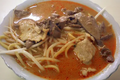A bowl of Xiamen shacha noodles in reddish-orange peanut satay broth with beef and bean sprouts