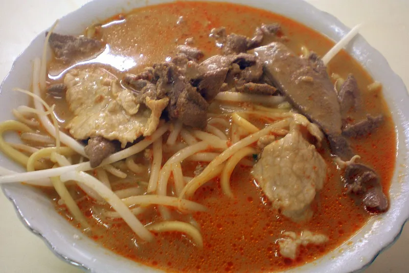 A bowl of Xiamen shacha noodles in reddish-orange peanut satay broth with beef and bean sprouts