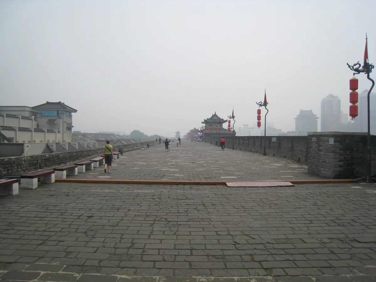Cyclists and pedestrians on the wide stone path atop Xi'an's ancient city wall, with a pagoda gate tower and the modern skyline visible through the haze