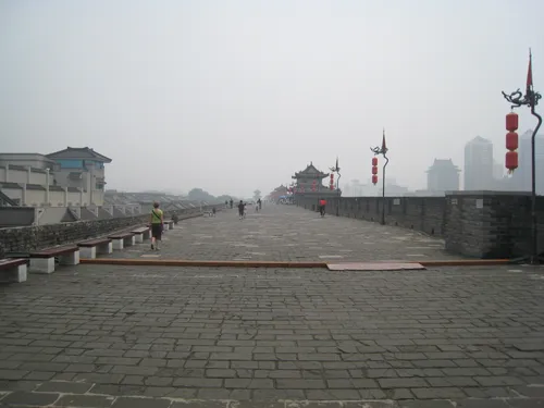 Cyclists and pedestrians on the wide stone path atop Xi'an's ancient city wall, with a pagoda gate tower and the modern skyline visible through the haze